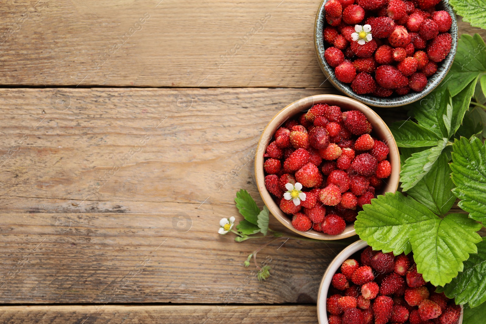 Photo of Fresh wild strawberries in bowls and leaves on wooden table, flat lay. Space for text