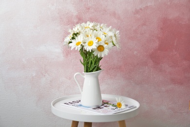 Jug with beautiful chamomile flowers on table against color background