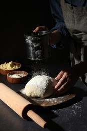 Woman sprinkling flour over dough at black table, closeup