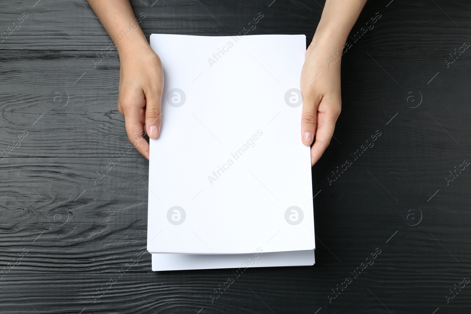 Photo of Woman holding blank paper sheets for brochure at black wooden table, top view. Mock up