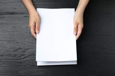 Woman holding blank paper sheets for brochure at black wooden table, top view. Mock up