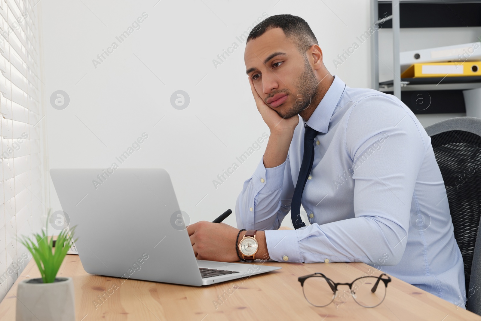 Photo of Puzzled young intern using laptop at table in modern office. First work day