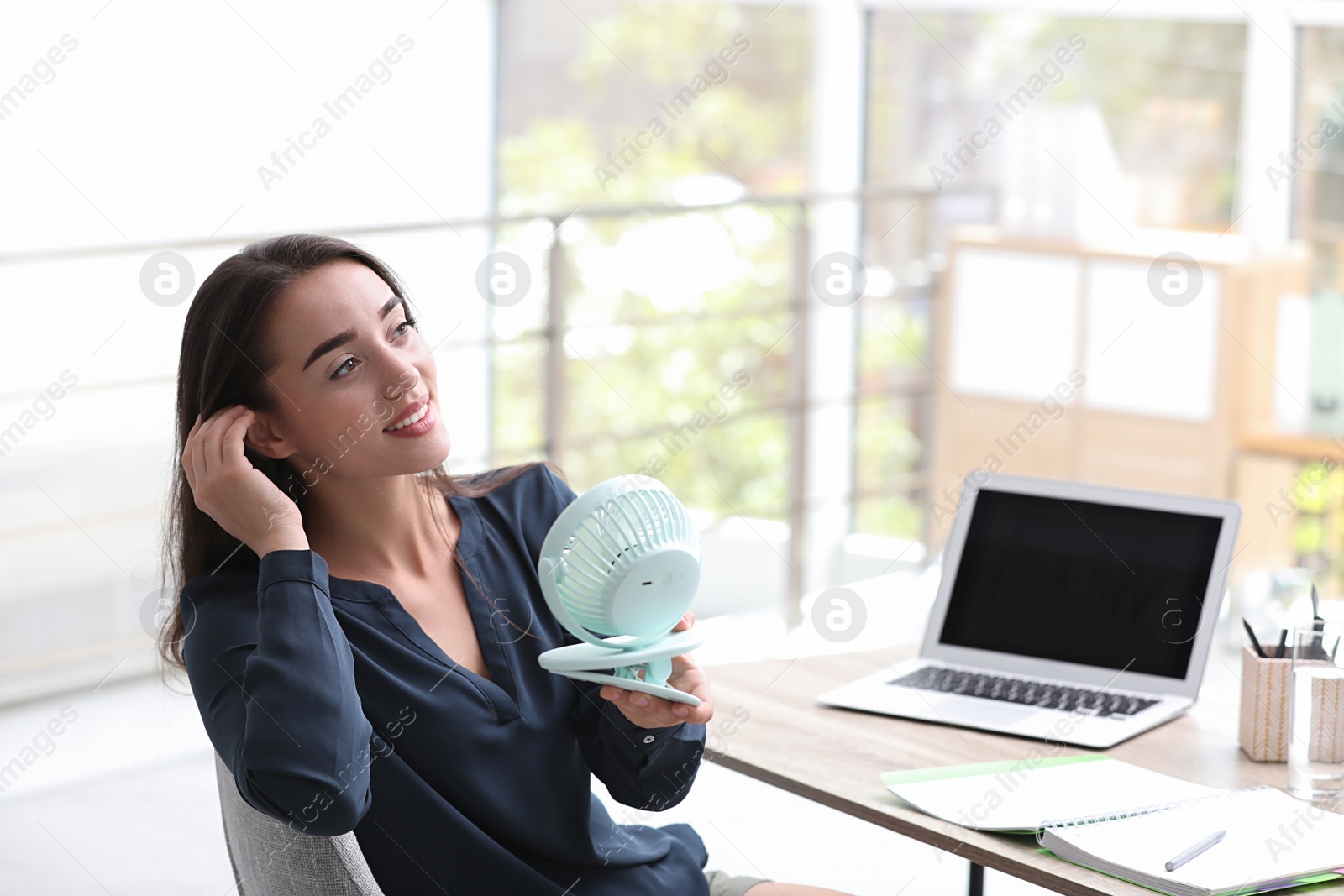 Photo of Young woman enjoying air flow from portable fan at workplace, space for text. Summer heat