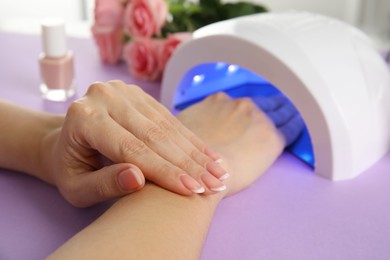 Photo of Woman using ultraviolet lamp to dry gel nail polish at violet table, closeup