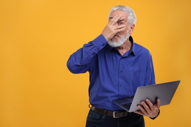Photo of Embarrassed senior man covering face with hand and holding laptop on orange background. Space for text