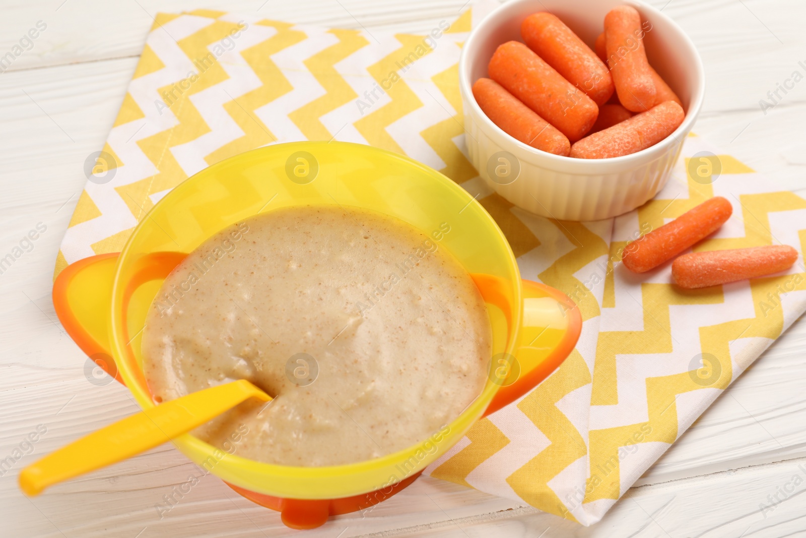 Photo of Baby food. Puree in bowl and small carrots on white wooden table