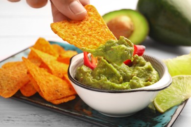 Photo of Woman dipping delicious nachos chip into guacamole at white wooden table, closeup