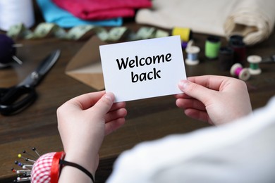 Image of Seamstress holding paper note with phrase Welcome Back at table in atelier, closeup