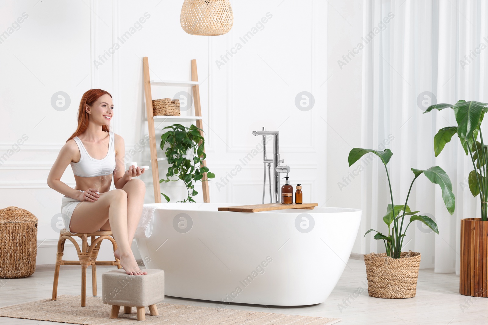 Photo of Beautiful young woman applying body cream onto legs in bathroom, space for text