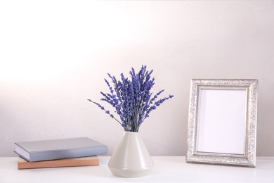 Photo of Bouquet of beautiful preserved lavender flowers, notebooks and blank photo frame on white table near beige wall