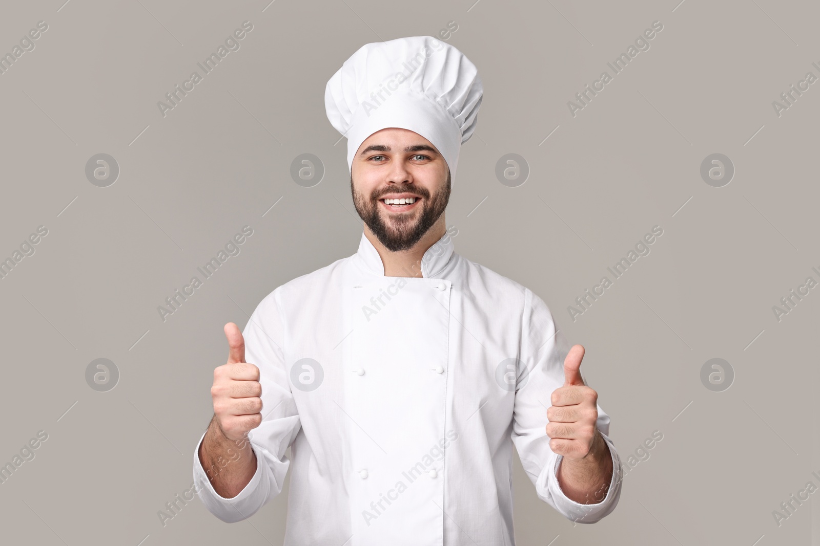 Photo of Happy young chef in uniform showing thumbs up on grey background