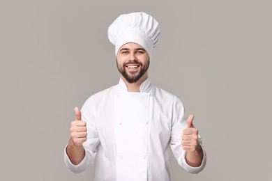 Photo of Happy young chef in uniform showing thumbs up on grey background