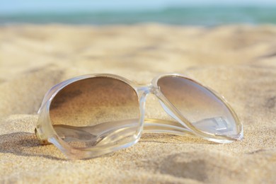 Photo of Stylish sunglasses on sandy beach near sea, closeup