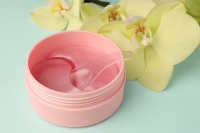 Photo of Jar of under eye patches with spoon and beautiful flowers on turquoise background, closeup. Cosmetic product