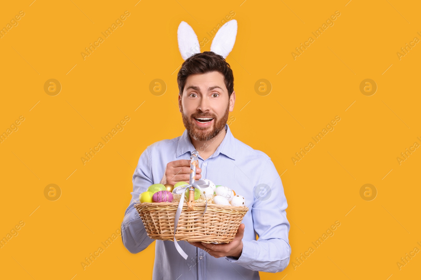 Photo of Portrait of happy man in cute bunny ears headband holding wicker basket with Easter eggs on orange background