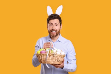 Portrait of happy man in cute bunny ears headband holding wicker basket with Easter eggs on orange background