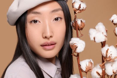 Beautiful young Asian woman with cotton branches on beige background, closeup