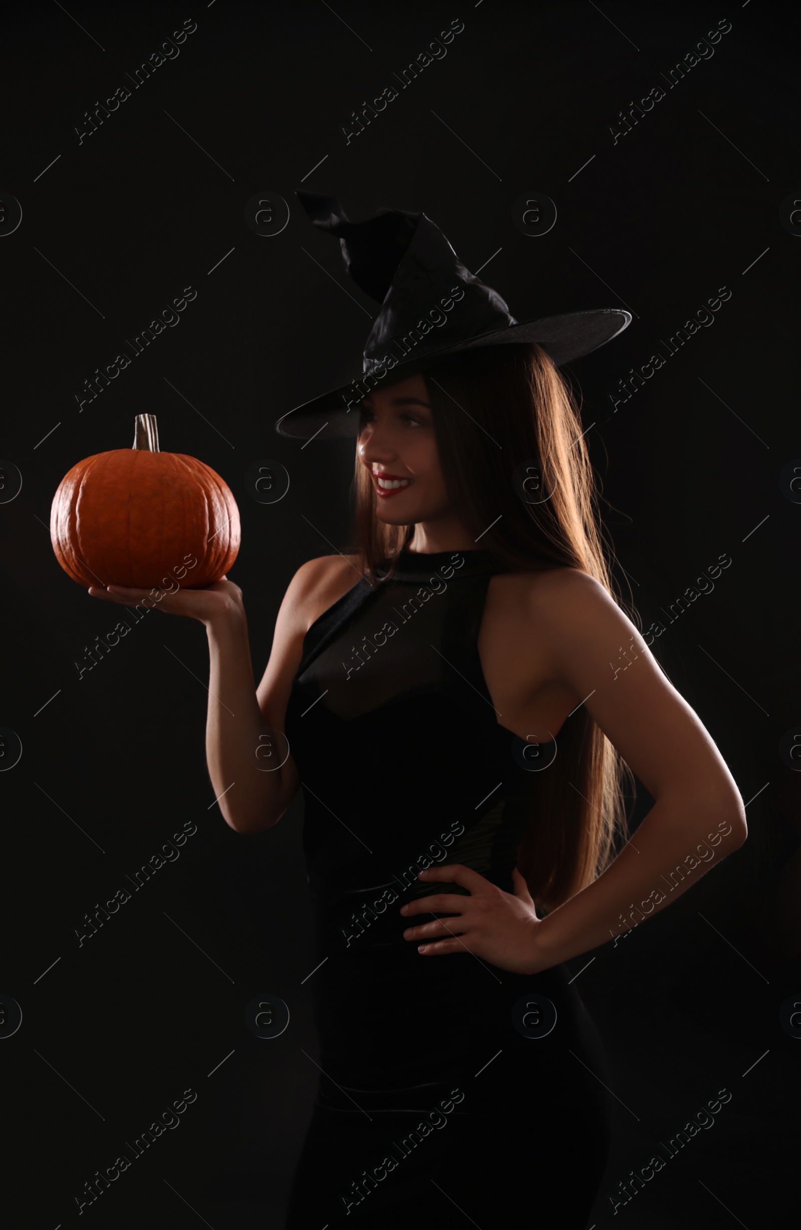 Photo of Young woman wearing witch costume with pumpkin on black background. Halloween party