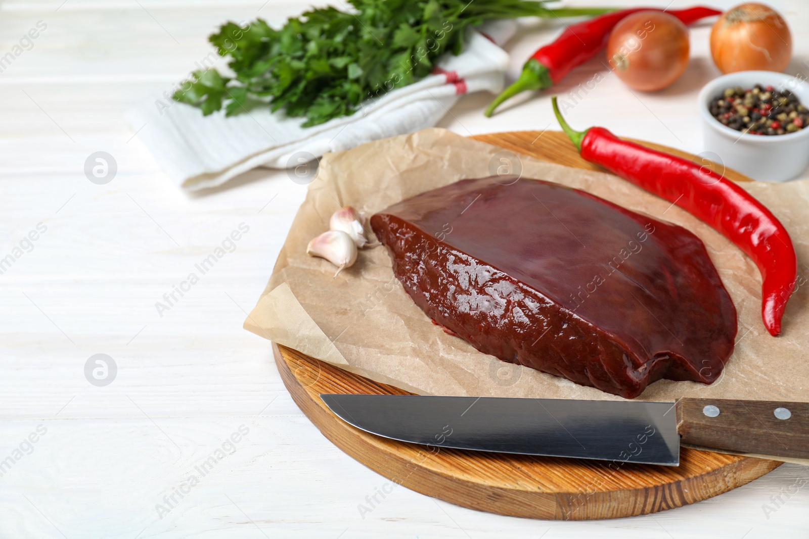 Photo of Piece of raw beef liver with chili pepper, knife and garlic on white wooden table, space for text