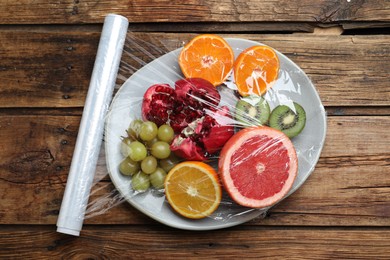 Photo of Plate of fresh fruits with plastic food wrap on wooden table, flat lay
