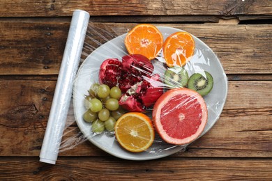 Plate of fresh fruits with plastic food wrap on wooden table, flat lay