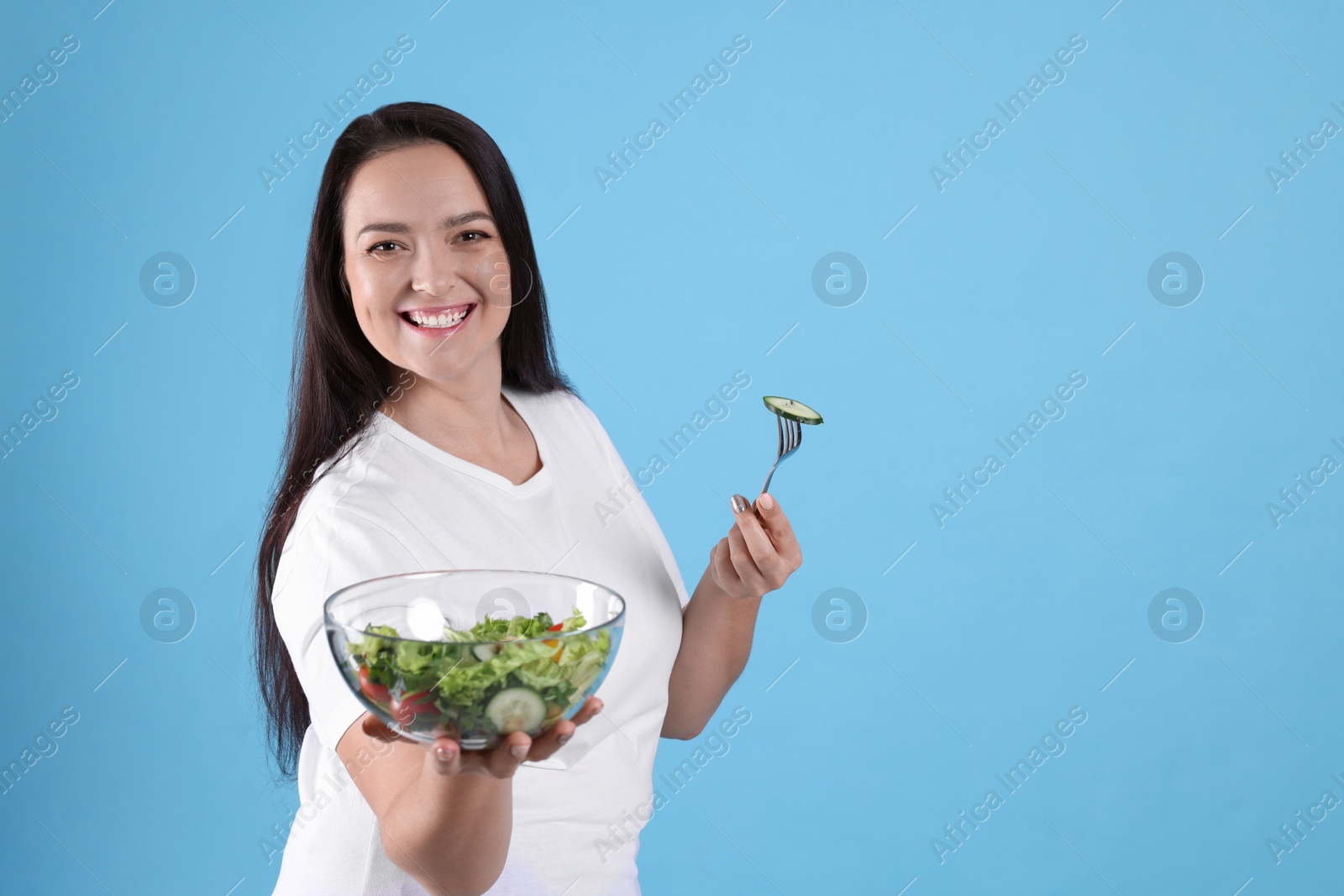 Photo of Beautiful overweight woman eating salad on light blue background, space for text. Healthy diet