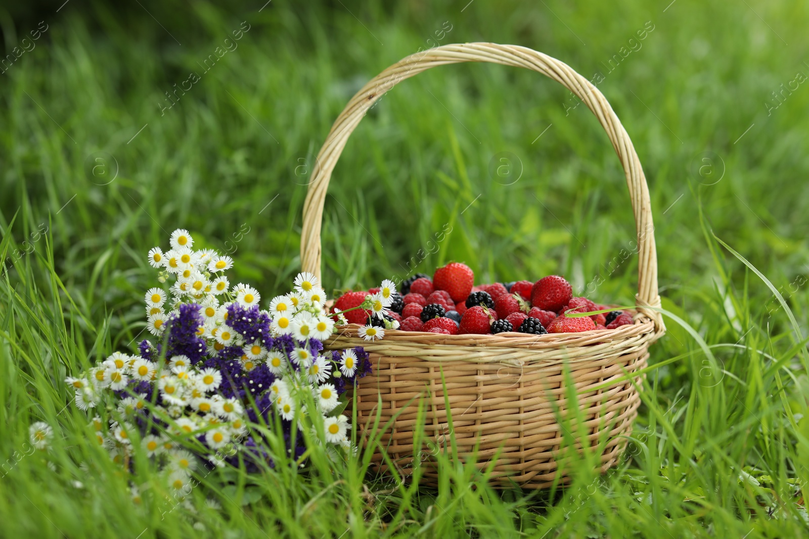 Photo of Wicker basket with different fresh ripe berries and beautiful flowers in green grass outdoors