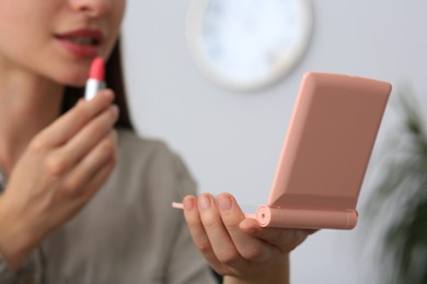 Photo of Young woman with cosmetic pocket mirror applying lipstick indoors, closeup
