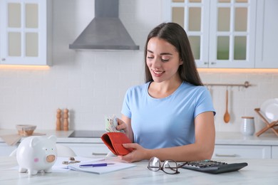 Young woman counting money at table in kitchen