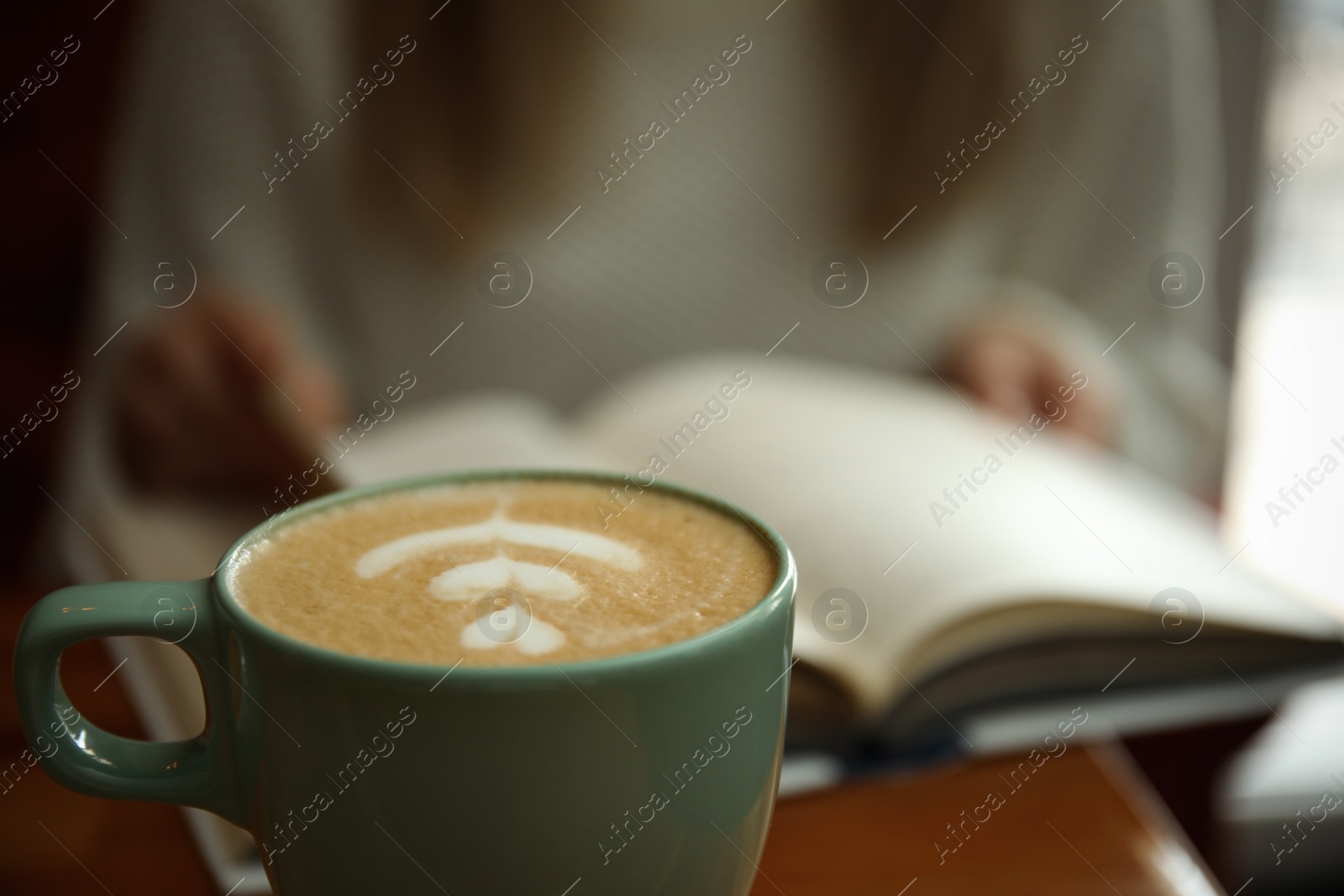 Photo of Woman with coffee reading book indoors, focus on cup