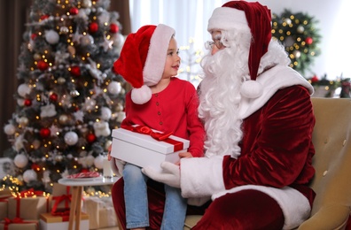 Photo of Santa Claus giving present to little girl in room decorated for Christmas