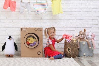 Little girl putting laundry into basket near toy cardboard washing machine indoors