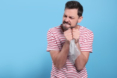 Angry man popping bubble wrap on light blue background, space for text. Stress relief