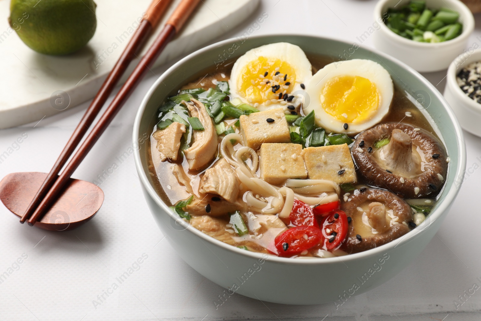 Photo of Noodle soup. Bowl of delicious ramen, ingredients and chopsticks on white tiled table, closeup