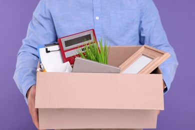 Unemployed man with box of personal office belongings on purple background, closeup
