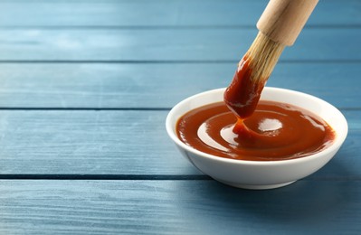 Photo of Tasty barbeque sauce in bowl and brush on blue wooden table, closeup. Space for text