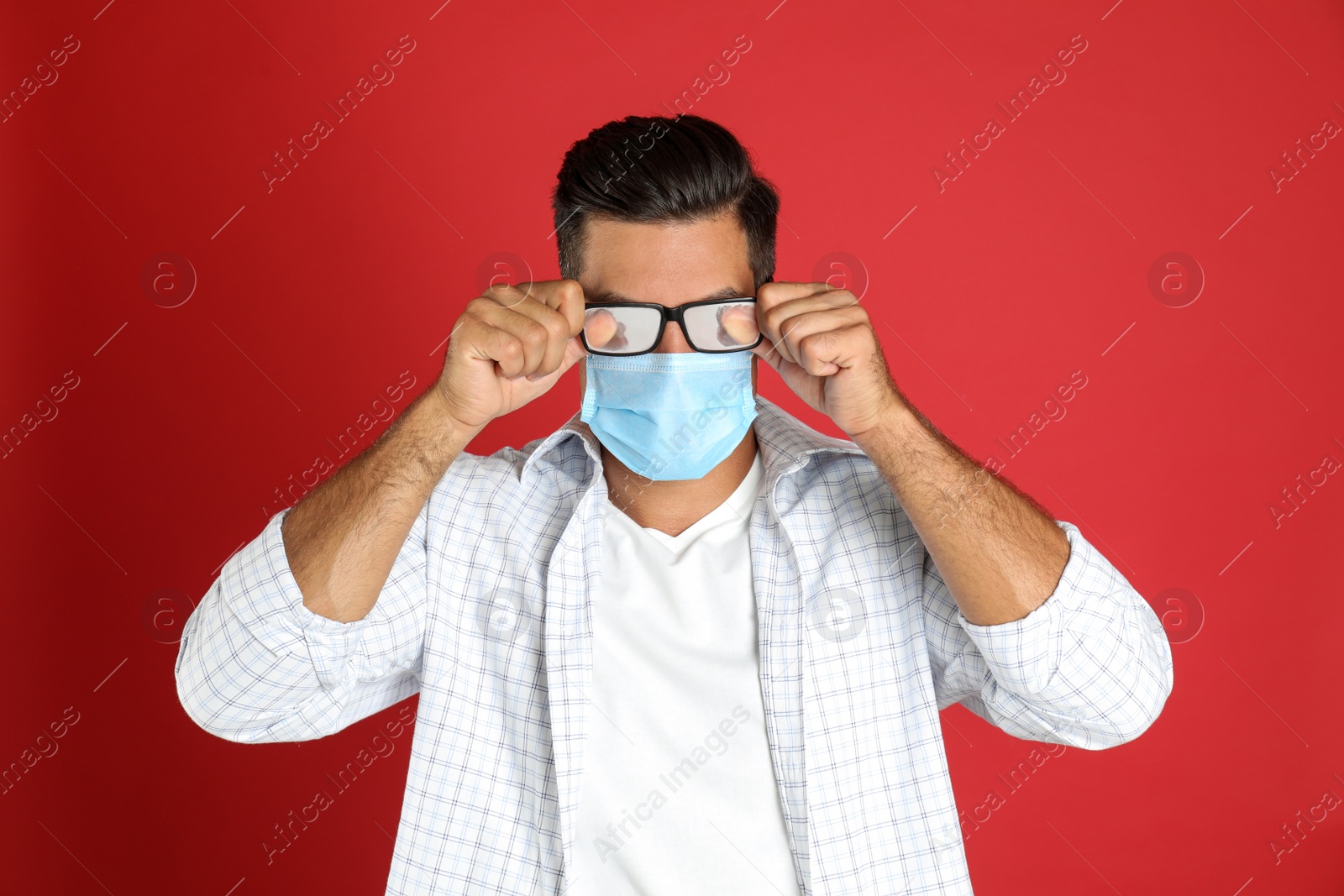 Photo of Man wiping foggy glasses caused by wearing medical mask on red background