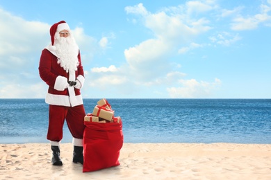 Santa Claus near sack of gifts on sandy beach. Space for text