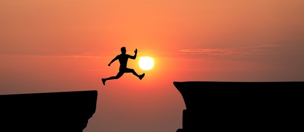 Image of Concept of reaching life and business goals. Silhouette of man jumping over chasm at sunrise