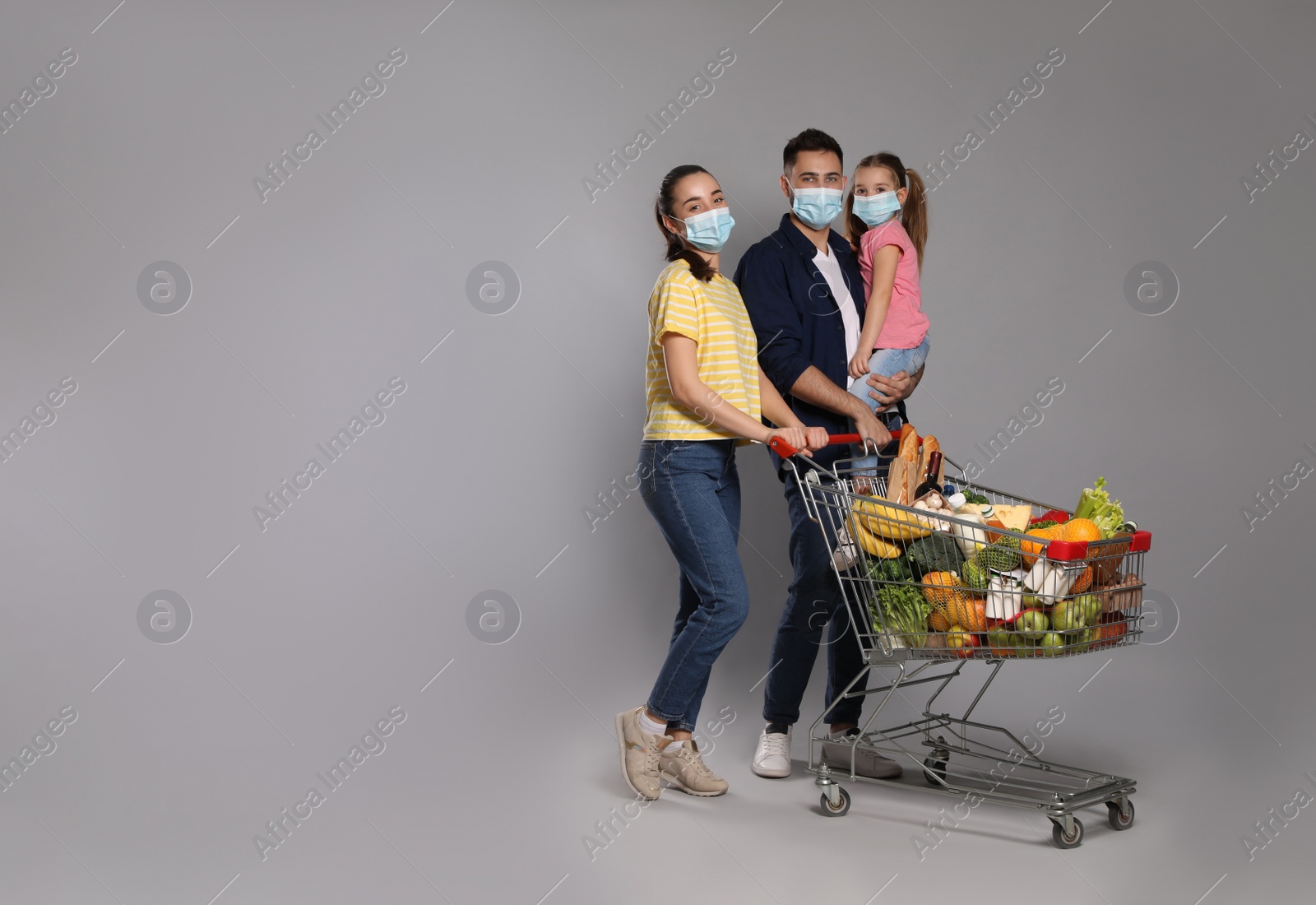 Photo of Family with protective masks and shopping cart full of groceries on light grey background