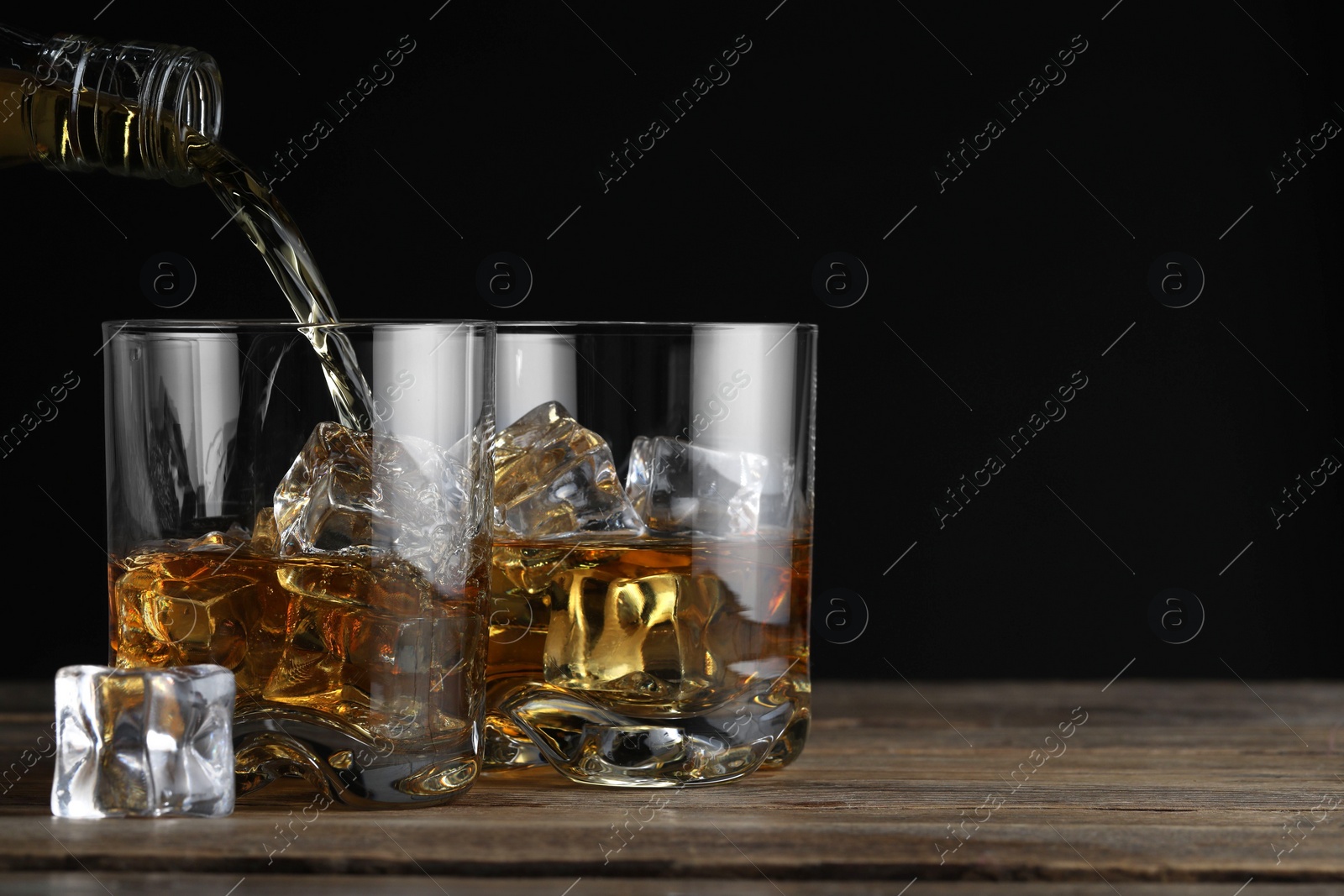 Photo of Pouring whiskey into glass with ice cubes at wooden table against black background, space for text