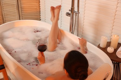 Photo of Woman with glass of wine taking bath in tub with foam and rose petals indoors, back view