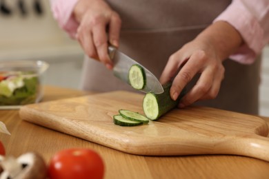Photo of Woman cutting cucumber at wooden table in kitchen, closeup