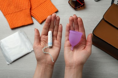 Woman holding violet menstrual cup and tampon over white wooden table, top view
