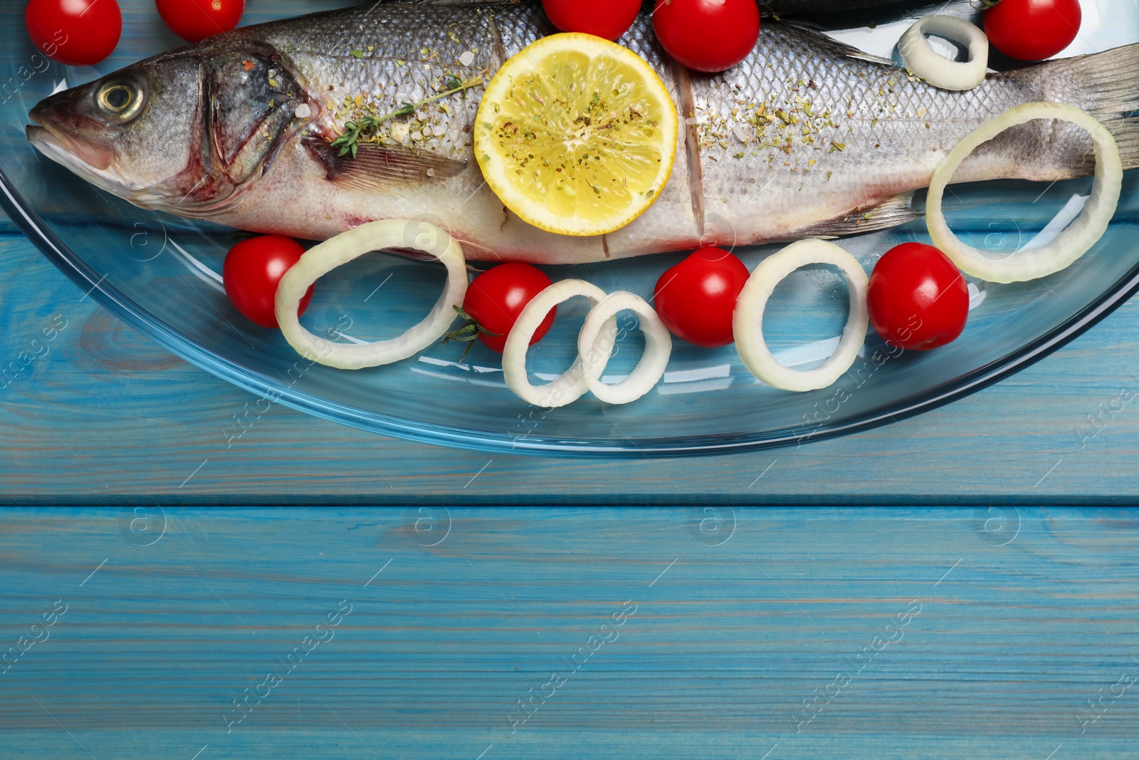 Photo of Glass baking tray with sea bass fish and ingredients on light blue wooden table, top view. Space for text