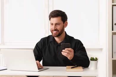 Smiling man with smartphone using laptop at table in office