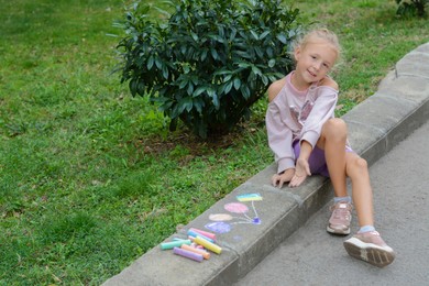 Photo of Little child sitting near drawn balloons and ukrainian flag with chalk on curb outdoors, space for text