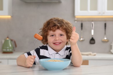 Cute little boy holding fork with sausage and showing thumbs up at table in kitchen