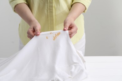 Photo of Woman holding shirt with stain at white table against light grey background, closeup
