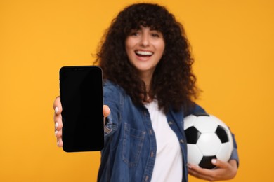 Photo of Happy fan holding soccer ball and showing smartphone on yellow background, selective focus